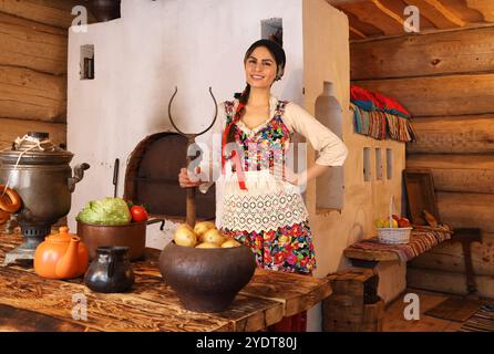 Eine junge Frau in traditioneller russischer Kleidung steht am Tisch mit Gemüse in einer typischen russischen Holzhütte Stockfoto