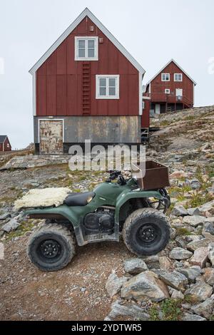 Ein grünes Quad oder ATV steht vor roten, holzverkleideten Häusern auf einem felsigen Hügel, der abgelegenen Arctic Inuit Siedlung Ittoqqortoormiit, Scoresbysund oder SC Stockfoto