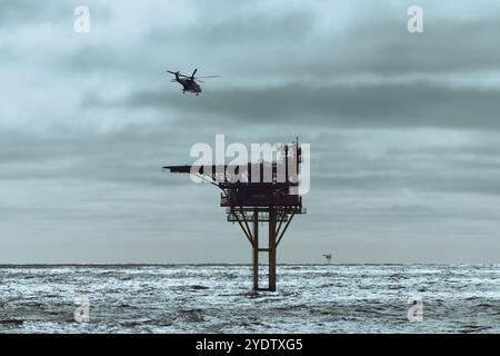 Hubschrauber fliegt zur Landung auf einer Offshore-Bohrinsel im Meer, mit Wolken am Himmel und blauem Meer. Stockfoto
