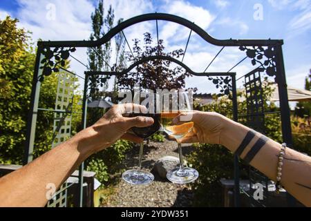 Ein paar Hände, die vor einem Gartentor des Weinguts rot- und Weißweingläser rösten und anfeuern, im Hintergrund verkostete Restaurant-Terrasse Stockfoto