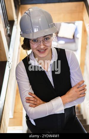 Junge attraktive kaukasische lächelnde Frau mit überkreuzten Armen posiert auf der Bautreppe, während sie einen grauen Schutzhelm und eine Schutzbrille trägt Stockfoto