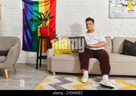 Eine queere Person sitzt bequem auf einem Sofa und arbeitet an einem Laptop, umgeben von farbenfroher Einrichtung. Stockfoto