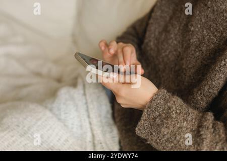 Nahaufnahme einer jungen Frau in einem gestrickten Pullover, die auf einem Sofa unter einer Decke sitzt und ihr Handy benutzt Stockfoto