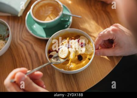 Blick von oben auf einen Mann, der eine Mango-Joghurt-Smoothie-Schüssel mit Banane, Mandel, Preiselbeeren, Kokos und einem Cappuccino-Kaffee isst Stockfoto
