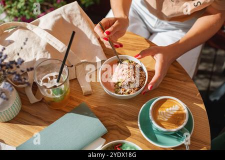 Blick von oben auf eine Frau, die an einem sonnigen Tag eine Joghurt-Smoothie-Schüssel mit Müsli, Kürbiskernen, Kokos und einer Tasse Kaffee isst Stockfoto