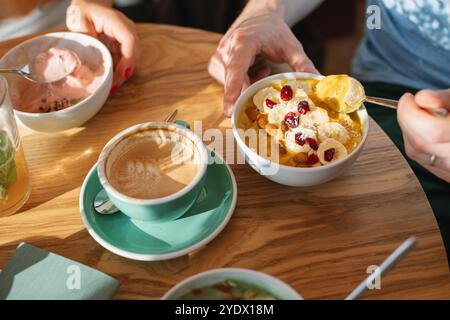 Blick von oben auf zwei Personen, die Jogurt Smoothie Bowls mit Banane, getrockneten Preiselbeeren und Kokos und einen Kaffee auf einer Terrasse in der Sommersonne genießen Stockfoto