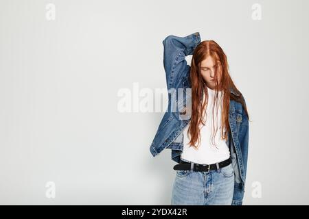 Ein Teenager mit auffälligen langen roten Haaren posiert selbstbewusst in einem Denim-Outfit und zeigt künstlerisches Flair. Stockfoto