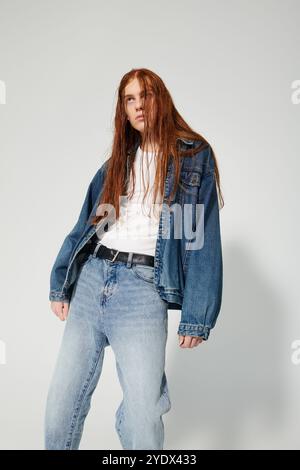 Ein Teenager mit auffälligen langen roten Haaren posiert selbstbewusst in einem Denim-Outfit vor StudioKulisse. Stockfoto