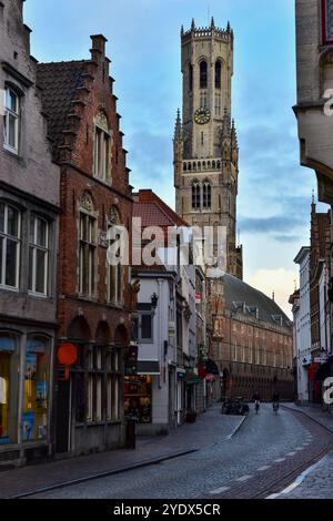 Glockenturm von Brügge, mittelalterliche Backsteinbauten und zwei Radfahrer kurz nach Sonnenaufgang auf der leeren Wollestraat in Brügge, Belgien Stockfoto