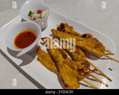 Gegrillte Hähnchenspieße, serviert mit Saucen, serviert auf einem langen weißen Teller und auf einem Tisch. Thailändische oder asiatische Küche. Stockfoto