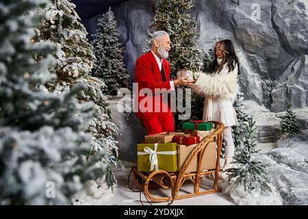 Ein fröhlicher Weihnachtsmann in Rot schenkt einer stilvollen Frau unter schneebedeckten Bäumen ein umhülltes Geschenk. Stockfoto