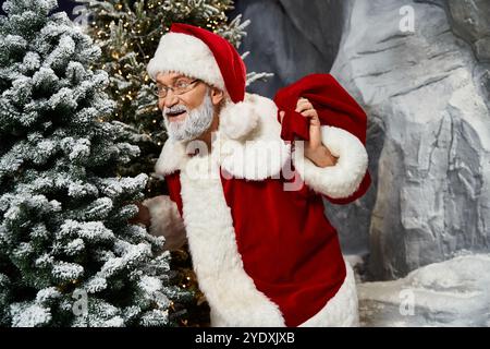 Eine fröhliche Figur, die als Weihnachtsmann gekleidet ist, bereitet Geschenke vor, während sie in der Nähe von schneebedeckten Bäumen steht. Stockfoto