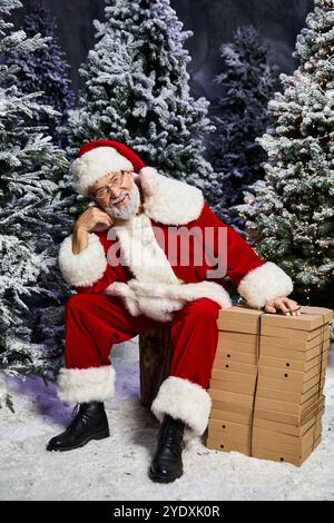 Ein fröhlicher Weihnachtsmann sitzt neben gestapelten Geschenken, umgeben von einem Winterwunderland aus Schnee. Stockfoto