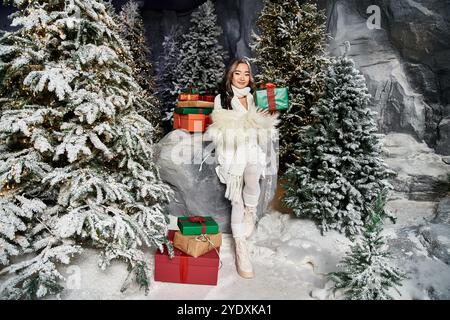 Ein junger Mensch hält ein wunderschön verpacktes Geschenk, umgeben von festlichen Dekorationen und Kiefern. Stockfoto
