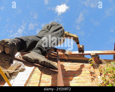 Beine eines Mannes mit den Sohlen seiner Stiefel, die von unten sichtbar sind, auf einer eisernen Leiter in der Nähe einer Ziegelwand stehen und Holzbalken unter dem Dach malen Stockfoto