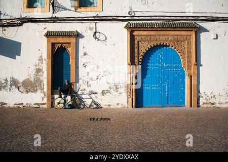 Essaouiras zeitloser Charme. Ein Mann mit seinem Fahrrad, der sich in der Abendsonne vor dem Hintergrund der traditionellen marokkanischen Architektur entspannt. Stockfoto