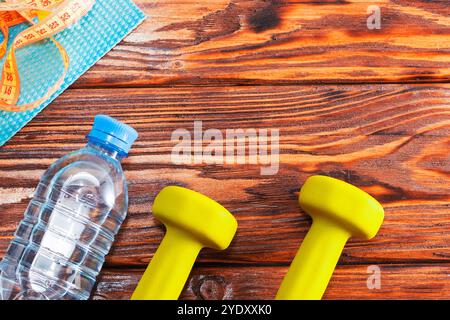 Auf einem Holztisch befinden sich zwei hellgelbe Kurzhanteln, eine Flasche Wasser gefüllt mit erfrischender Flüssigkeit und ein Maßband, das ordentlich platziert ist Stockfoto