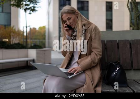 Eine Geschäftsfrau sitzt auf einer Bank vor einem Business Center, arbeitet an ihrem Laptop und spricht mit ihrem Telefon Stockfoto