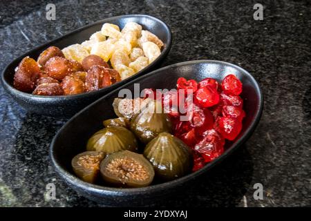 Zwei schwarze Wüstenschalen, die jeweils mit zwei verschiedenen kandierten Früchten gefüllt sind, auf einer Oberfläche aus schwarzem Granit. Stockfoto