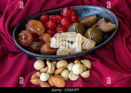 Schwarze Desert-Schüssel gefüllt mit einem Medley aus konservierten Früchten, umgeben von verschiedenen Nüssen auf weichem rotem Material Stockfoto