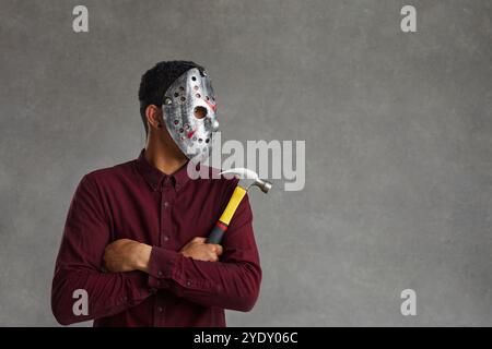 Schwarzer Mann in gruseliger Maske, der Hammer hält und auf grauen Hintergrund blickt Stockfoto