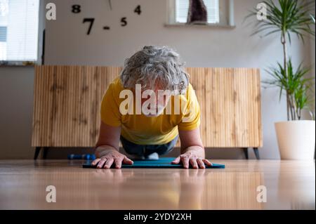 Ein konzentrierter Seniorenmann mit lockigen grauen Haaren hält eine Plank-Position auf einer Yogamatte und zeigt damit Belastbarkeit und Engagement für aktiv zu bleiben. Stockfoto