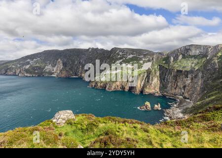 Die Klippen der Slieve League in Irland überblicken eine kleine Bucht mit einsamen Felsen, umgeben von grünen Hügeln und dem Atlantischen Ozean unter einem teilweise bewölkten Himmel Stockfoto