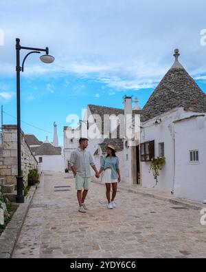 Ein Paar genießt einen gemütlichen Spaziergang auf einer Kopfsteinpflasterstraße in Alberobello Apulien, Italien. Umgeben von traditionellen Trullihäusern mit konischen Dächern teilen sie einen freudigen Moment unter dem weiten Himmel. Stockfoto