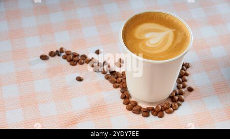 Kaffeepapierbecher zum Mitnehmen mit Kaffeebohnen auf kariertem Hintergrund. Milchschaumherz Stockfoto