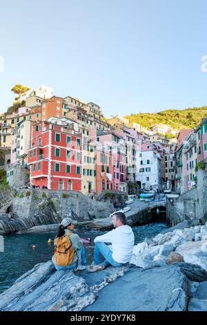 Zwei Freunde sitzen auf zerklüfteten Felsen und genießen die atemberaubende Aussicht auf farbenfrohe Häuser am Ufer von Cinque Terre und genießen die warme Sonne an einem wunderschönen Tag. Riomaggiore in Cinque Terre, Italien Stockfoto
