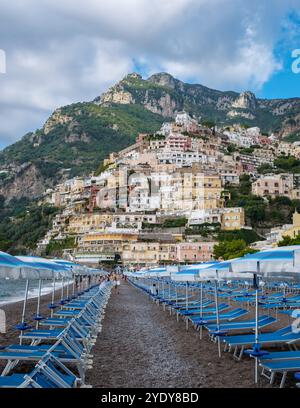 Besucher schlendern entlang eines Sandstrandes, der von pulsierenden Sonnenliegen gesäumt ist, während bezaubernde Häuser an den Klippen die Amalfiküste schmücken. Positano auf Italien Amalfi CoA Stockfoto