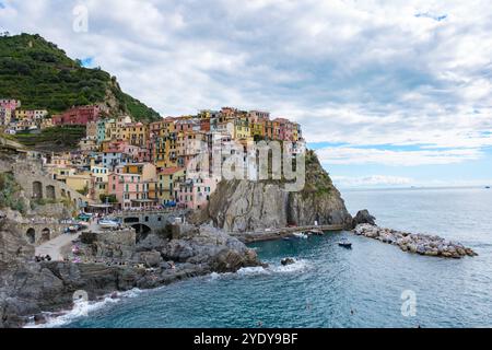 Eingebettet an der felsigen Küste, sind die farbenfrohen Gebäude von Manarola vom azurblauen Meer eingerahmt, was eine malerische Aussicht bietet. An einem ruhigen Nachmittag genießen die Besucher Freizeitaktivitäten inmitten der atemberaubenden Naturschönheit. Stockfoto