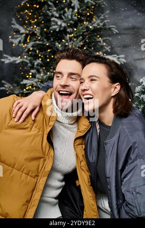 Ein glückliches Paar umarmt sich in gemütlichen Jacken, umgeben von schneebedeckten Bäumen, um den Weihnachtsgeist zu feiern. Stockfoto
