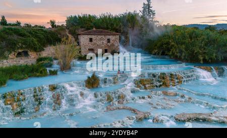 Warmer Dampf steigt auf, während Besucher sich im natürlichen Thermalwasser von Saturnia entspannen, umgeben von atemberaubenden Landschaften. Das alte Steingebäude verleiht diesem malerischen abendlichen Kurzurlaub in der Toskana Charme. Stockfoto