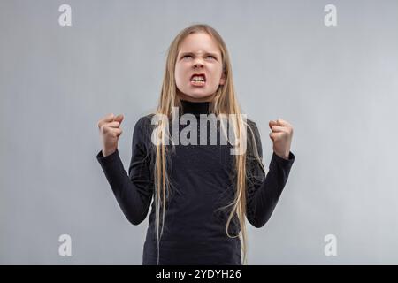 Ein junges Mädchen mit blonden Haaren und einem schwarzen Hemd hebt ihre Fäuste und zeigt ihre Zähne vor grauem Hintergrund, mit Trotz und s Stockfoto