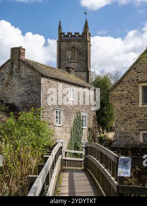 Eine Fußgängerbrücke überquert den Nunney Brook neben der All Saints Church im Somerset Village Nunney. Stockfoto