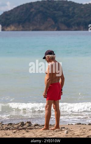 Ein älterer Mann mit roten Shorts und einer Baseballmütze, der an einem Sandstrand steht und allein aufs Meer blickt. Stockfoto