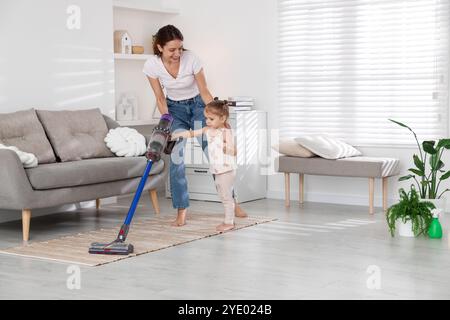 Die lächelnde junge Frau und ihre Tochter putzen zu Hause den Boden mit schnurlosem Staubsauger Stockfoto