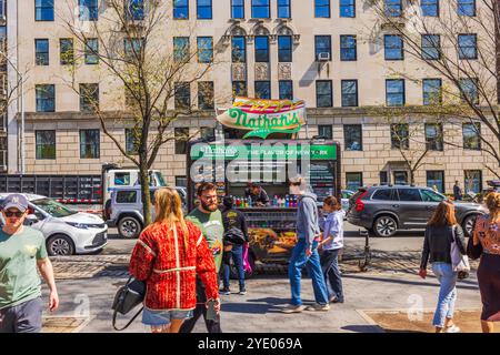 Menschen, die an sonnigen Tagen an Nathan's berühmtem Hot Dog Stand im Central Park vorbeilaufen, mit einem großen Gebäude im Hintergrund. New York. USA. Stockfoto