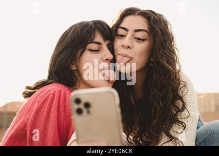 Authentischer Moment, in dem verschiedene Freunde der Generation Z Selfie im Freien machen. Junge Frauen mit lockigen Haaren und Zahnspangen, die lustige Gesichter an der Smartphone-Kamera machen, echt Stockfoto