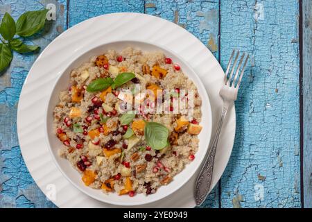 Quinoa-Salat mit geröstetem Kürbis, frischem Apfel, Granatapfelkernen, Pekannuss, getrockneten Preiselbeeren und Basilikumblättern in einer weißen Schüssel mit Gabel auf blauem Holztablett Stockfoto