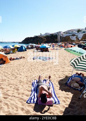 Sonnenanbeter entspannen sich auf dem goldenen Sand von Albufeira, ein ruhiger Tag unter pulsierenden Sonnenschirmen mit malerischen Klippen im Hintergrund Stockfoto