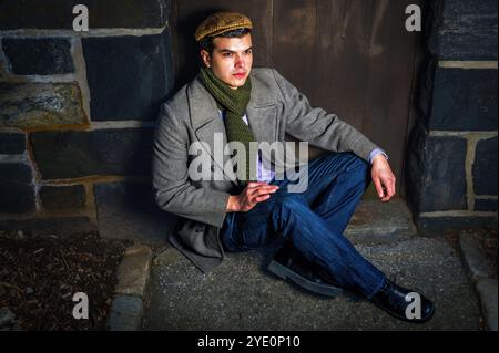 Ein Mann sitzt an einer Steinmauer, gekleidet mit Wollmantel, Schal und Mütze. Die Szene hat eine stimmungsvolle Atmosphäre mit kontrastierender Beleuchtung, die das t Stockfoto