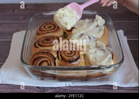 Eine Frau verteilt mit einem Küchenspachtel Creme auf gebackene Zimtbrötchen in transparenter Form auf dem Tisch Stockfoto