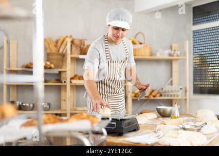 Junger positiver männlicher Bäcker in Schürze, Teig schneiden und portionierte Stücke auf dem Tisch wiegen, Prozess der Brotzubereitung in der Küche Stockfoto