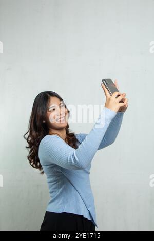 Eine Frau in einem hellblauen Kleid macht ein Selfie mit ihrem Handy, die Frau hat lange Haare und sieht fröhlich aus, wenn sie auf weißem Hintergrund steht. Stockfoto
