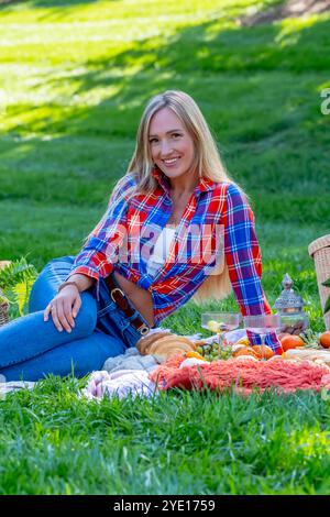 Ein atemberaubendes europäisches Model, gekleidet in gemütlichen Herbstschichten, genießt an einem frischen Herbsttag ein malerisches Picknick im Freien. Goldene Blätter umgeben sie während sie Stockfoto