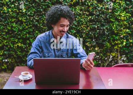 Ein junger asiatischer Mann mit lockigen Haaren, der ein Jeanshemd trägt, lächelt, während er mit Laptop und Kaffee an einem Tisch sitzt und ein Smartpho hält Stockfoto