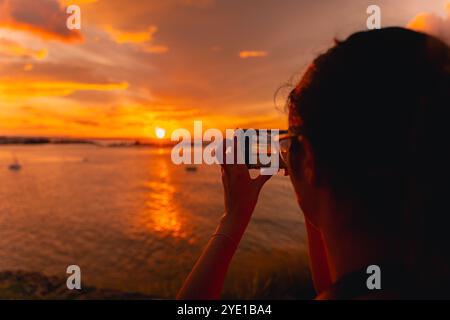 Eine Person fängt den lebhaften Sonnenuntergang über Einem ruhigen Meer mit ihrem Smartphone ein, das sich vor dem feurigen Orange und dem Gelben Himmel in der Nähe von El Morro, Puerto, befindet Stockfoto