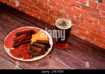 Ein Glas halbdunkles Bier und ein großer Teller mit einer Auswahl an Fleischgerichten, Käse und Roggencroutons auf einem dunklen Holztisch. Nahaufnahme. Stockfoto